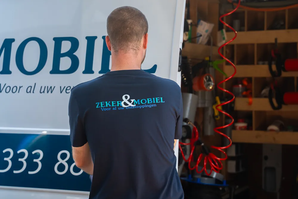 Monteur van Zeker & Mobiel in werkkleding met ruglogo en gereedschap, klaar om een verstopping te verhelpen