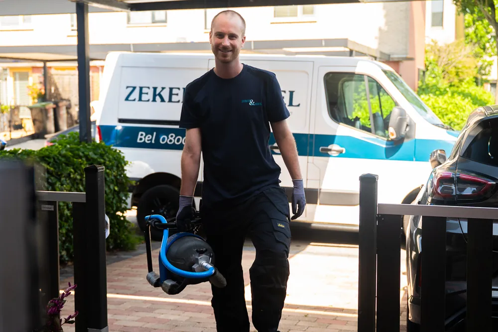 Monteur van ontstoppingsdienst Zeker & Mobiel loopt met de ontstoppingsmachine naar de voordeur om een riool te ontstoppen, de bus staat achter hem