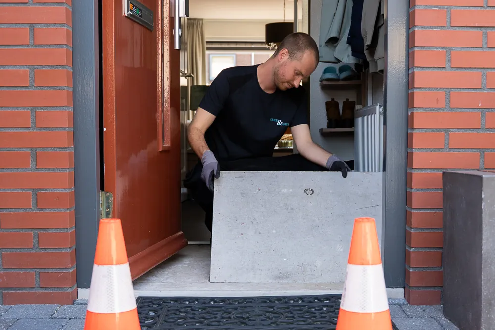 Riool ontstoppen: monteur van Zeker & Mobiel tilt het deksel van de put in de gang bij de voordeur, met twee pionnen op de stoep